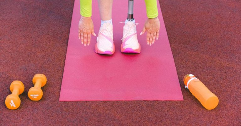 Exercise Motivation - Unrecognizable female athlete with leg prosthesis bending forward and stretching while trying ti touch ground during training on sports ground with dumbbells