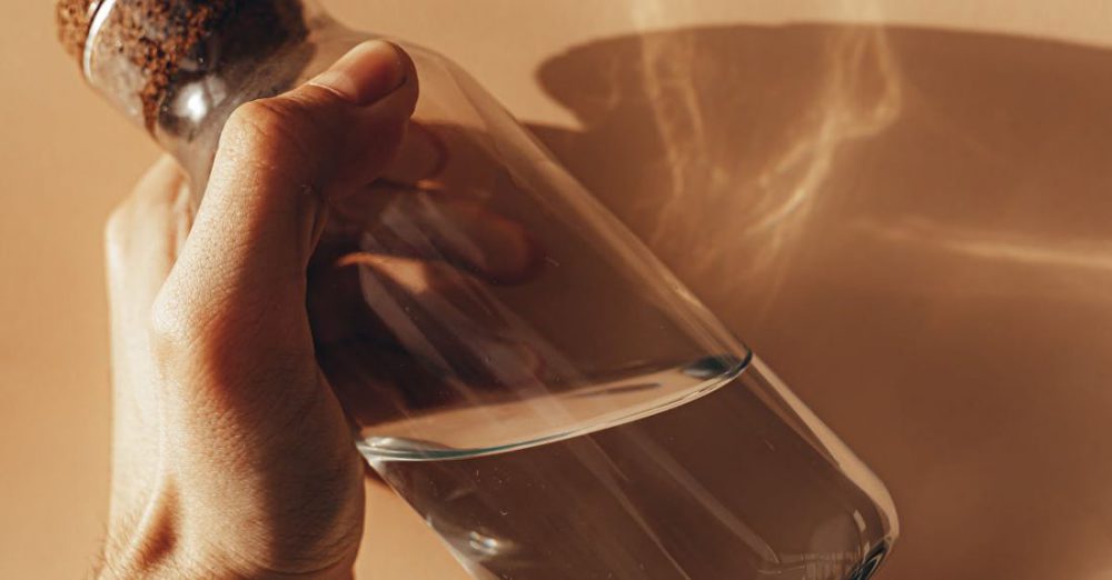 Detoxing - Crop anonymous male holding transparent glass bottle of water in hand against beige wall with shades during sunny day