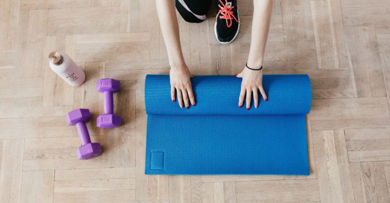 Yoga Accessories - Top view of anonymous female athlete in black leggings and sneakers unfolding blue mat for exercising on floor near dumbbells and water bottle in modern fitness center
