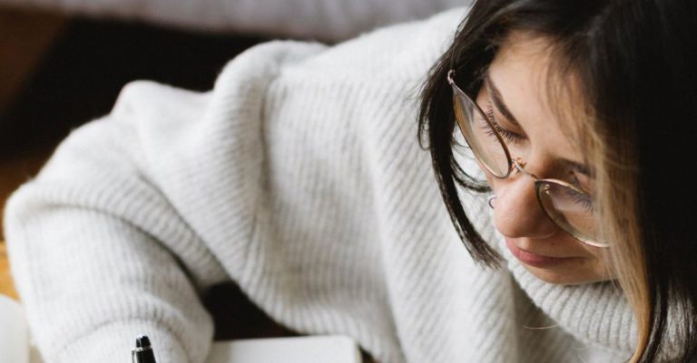 Sleep Schedule - From above of crop young female in warm clothes and eyeglasses writing plan for 2021 in notebook while sitting on floor near sleeping Akita Inu dog